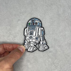 Könnte beinhalten: Gestickter Aufnäher mit dem ikonischen R2-D2-Droiden aus Star Wars. Der Aufnäher ist überwiegend weiß mit blauen und schwarzen Akzenten, mit detaillierten Stickereien und einem schwarzen Rand. Der Aufnäher ist etwa 7.6 cm hoch.
