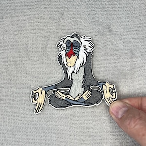 May include: Embroidered patch featuring a cartoon baboon in a seated yoga pose. The baboon has gray fur, a red nose, and a white beard. The patch is held by a hand against a gray background.