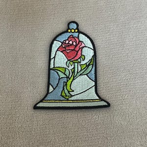 May include: A red rose with green leaves and stem is encased in a glass dome with a blue and white stained glass design. The dome is outlined in gold and black.