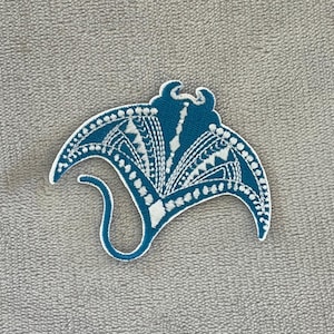 Peut inclure: Écusson brodé bleu sarcelle et blanc en forme de raie manta. Le patch présente des motifs blancs complexes et une bordure blanche. La raie manta a une queue incurvée et se détache sur un fond gris.