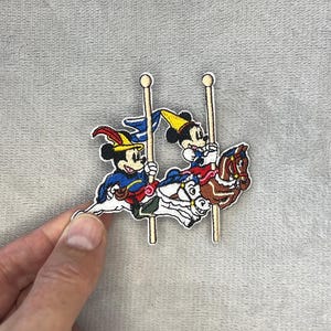 May include: Embroidered patch featuring Mickey Mouse and another character riding carousel horses. The characters are dressed in colorful outfits, with Mickey wearing a yellow hat. The patch has a white border and is set against a gray background.