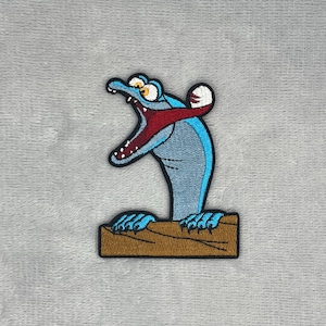 May include: An embroidered patch featuring a cartoon creature with light blue skin, a wide open mouth with a red interior, white teeth, and yellow eyes. The creature has blue claws resting on a brown rectangular log-like base with a black border.