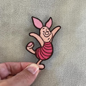 Może przedstawiać: Haftowana naszywka przedstawiająca Prosiaczka z Kubusia Puchatka. Naszywka przedstawia Prosiaczka w radosnej pozie, z wyciągniętymi ramionami i jedną zgiętą nogą. Projekt zawiera odcienie różu, brązu i czerni, z paskiem na ciele.