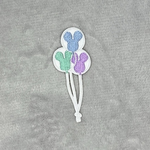 May include: Embroidered patch featuring three balloons shaped like a mouse head. The balloons are blue, green, and purple with white outlines and strings. The patch is on a grey, textured background.