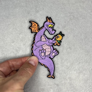 Könnte beinhalten: Gestickter Aufnäher mit einem lila Drachen mit orangefarbenen Flügeln und einem gelb-orangenen Vogel. Der Drache hat einen rosa Bauch und hält den Vogel. Der Aufnäher hat einen schwarzen Rand.