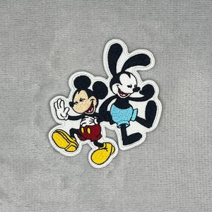 May include: An embroidered patch featuring Mickey Mouse and Oswald the Lucky Rabbit. Mickey wears red shorts and yellow shoes, while Oswald wears light blue shorts. Both characters are depicted in a dynamic pose with white outlines on a grey background.