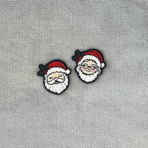 Puede incluir: Dos parches bordados con caras de Papá Noel. Cada parche tiene un gorro rojo con ribete blanco, una barba blanca y una expresión sonriente. Los parches están delineados en negro y tienen un pequeño acento de acebo verde.