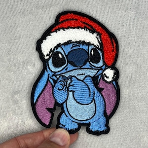 Puede incluir: Parche bordado con el alienígena azul Stitch de Lilo & Stitch, con un gorro de Papá Noel rojo y blanco. El parche tiene un borde negro y representa a Stitch con una expresión triste, ideal para manualidades navideñas.