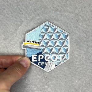 以下が含まれることがあります： EPCOTセンターのロゴが入った刺繍パッチ。幾何学的な球体、モノレール、白い文字で「EPCOT CENTER」の文字がデザインされています。パッチは、水色、グレー、白の配色です。