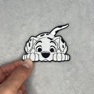 May include: Embroidered patch featuring a black and white Dalmatian puppy. The patch shows the puppy's face and paws, with black spots on a white background. The puppy's tail is raised. The patch is on a gray surface.