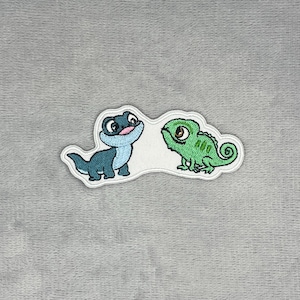May include: An embroidered patch featuring two cartoon creatures. On the left, a small blue lizard with a light blue belly and large eyes. On the right, a green chameleon with a curled tail and large brown eyes. The patch has a white border.