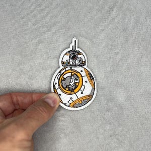May include: Embroidered patch of the BB-8 droid from Star Wars. The patch has a white border and shows BB-8 with orange, grey, and yellow details. The patch is held by a hand against a grey background.