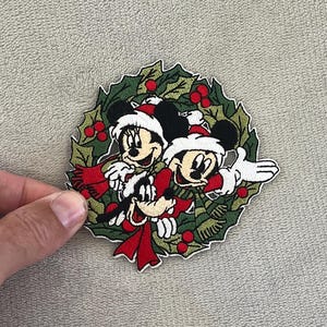 Peut inclure: Écusson brodé représentant Mickey Mouse, Minnie Mouse et Dingo dans une couronne de Noël. Les personnages portent des bonnets de Père Noël et des écharpes rouges, entourés de feuilles de houx vertes et de baies rouges. L'écusson est circulaire.