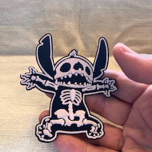 May include: A black and white embroidered patch depicting a cartoon character as a skeleton. The character has large ears and outstretched arms. The patch is held in a hand, showing the detail of the design.