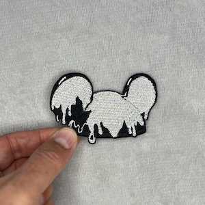 May include: Black and white embroidered patch in the shape of a mouse head with dripping white paint effect. The patch is held by a hand against a gray background.