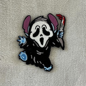 Peut inclure: Écusson brodé représentant un personnage de dessin animé en robe noire et masque blanc hurlant. Le personnage tient un couteau ensanglanté. L'écusson a des oreilles violettes et des accents bleus.