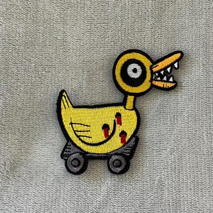 以下が含まれることがあります： 脅迫的な表情の黄色いアヒルの刺繍パッチ。アヒルは大きく丸い目、鋭い歯、赤いアクセントを持っています。4つの車輪に取り付けられており、ユニークで漫画のような外観になっています。