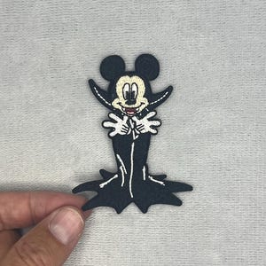 May include: An embroidered patch featuring a cartoon mouse character dressed as a vampire. The character has a cream-colored face, black ears, and is wearing a long black cape with a high collar, white gloves, and visible fangs against a dark background.