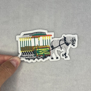 May include: An embroidered patch featuring a white horse pulling a green and yellow trolley car. The trolley has vertical green poles, a dark roof, and a decorative yellow scroll design on its side. The horse is depicted in white with black harness details.