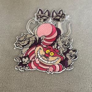 Op de afbeelding: Geborduurde patch met de Cheshire Cat uit Alice in Wonderland, met een grote grijns en roze en rode strepen. Rondom de kat staan andere kattenpersonages in tinten paars, grijs en roze.