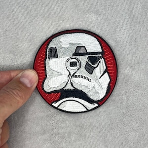 May include: Embroidered patch featuring a Stormtrooper helmet design. The patch is circular, with a red border framing the white and black helmet. The helmet has detailed stitching, capturing the iconic look from the film.