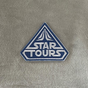Könnte beinhalten: Ein blau-weißer, gestickter Aufnäher in Dreiecksform. Der Aufnäher zeigt die Worte "STAR TOURS" in weißer Schrift mit einem stilisierten Design über dem Text. Der Aufnäher ist wahrscheinlich für Kleidung oder Accessoires.