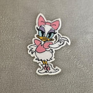 Puede incluir: Parche bordado para planchar con un pato de dibujos animados con un lazo rosa, un vestido rosa y zapatos blancos. El pato tiene ojos azules y un pico amarillo.