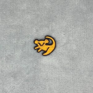 May include: A small embroidered patch featuring a stylised yellow lion cub silhouette outlined in dark brown thread. The design depicts a crouching lion cub with a curved tail, creating a circular shape. The embroidery has a textured finish.
