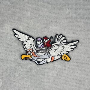 Può includere: Una toppa ricamata raffigurante un grande uccello bianco con becco giallo e occhiali arancioni, in volo mentre trasporta due piccole figure in un cestino. Una figura indossa un cappello rosso e l'altra un cappello viola.