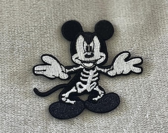 May include: Black and white embroidered patch featuring a cartoon mouse with a skeleton design. The mouse has large black ears, a black tail, and white hands and feet. The patch is likely for clothing or accessories.
