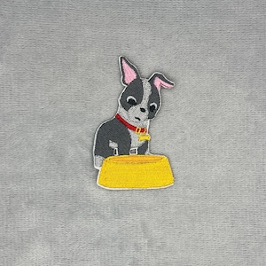 May include: An embroidered patch featuring a small gray and white dog with large ears and a pink inner ear detail. The dog wears a red collar with a yellow bone-shaped tag and is positioned behind a solid yellow bowl with a textured surface.