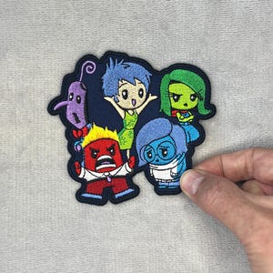May include: An embroidered patch featuring five colorful cartoon characters. The group includes a purple figure, a blue-haired character in a green dress, a green-skinned character, a red character with yellow hair, and a blue character in a sweater.