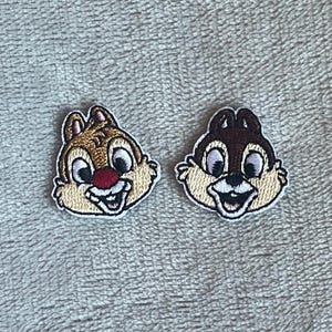 May include: Two embroidered patches featuring the faces of Chip and Dale, Disney cartoon chipmunks. Chip is on the left with a tan face, red nose, and brown hair. Dale is on the right with a brown face and black nose. Both have white teeth and are smiling.
