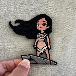 May include: Embroidered patch depicting a cartoon Native American woman with long black hair. She wears a light pink dress with white fringe and stands on a grey rock. The patch is designed for clothing or accessories.
