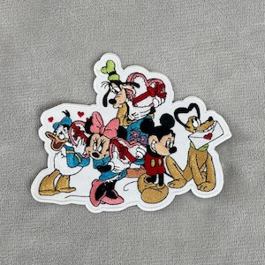 May include: An embroidered patch featuring Mickey Mouse, Minnie Mouse, Donald Duck, Goofy, and Pluto. The characters are all smiling and holding hearts or letters. The patch is white with a red, blue, yellow, and green colour scheme.