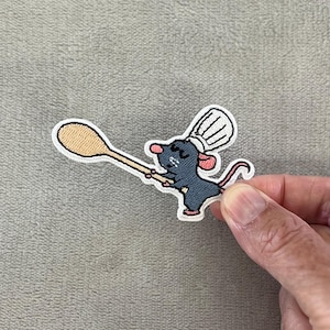 Può includere: Toppa ricamata raffigurante un topo chef dei cartoni animati con un cappello da cuoco bianco e un cucchiaio di legno. Il topo è grigio con orecchie rosa e una coda rossa. La toppa ha un bordo bianco.