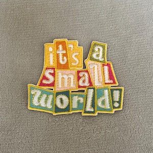 Może przedstawiać: Haftowana naszywka z napisem "it's a small world!" wykonanym białą nicią. Litery umieszczone są na tle wielokolorowych, nieregularnych prostokątnych kształtów w odcieniach żółtego, pomarańczowego, różowego, czerwonego, turkusowego i zielonego.