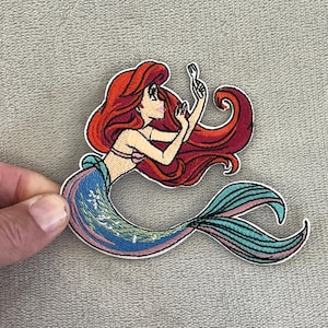May include: Embroidered patch of a mermaid with red hair, a pink top, and a blue and teal tail. The mermaid is holding a fork. The patch has a white border.