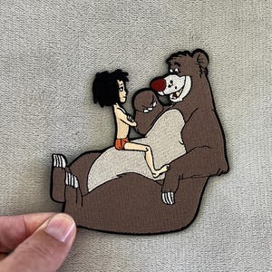 May include: A brown and tan embroidered patch featuring a cartoon bear and a boy. The bear has a red nose and is smiling. The boy is sitting on the bear's back. The patch has a black border.