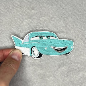 Könnte beinhalten: Gestickter Aufnäher mit einem türkisfarbenen Cartoon-Auto mit lächelndem Gesicht, weißen Akzenten und schwarzen Reifen. Das Auto hat eine Finne und grüne Augen. Der Aufnäher wird vor einem grauen, strukturierten Hintergrund gehalten.