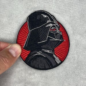 May include: Embroidered patch featuring Darth Vader's profile in black, set against a red circular background with radiating lines. The patch is held by a hand, showcasing the detailed stitching and design.