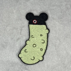 May include: Embroidered patch featuring a cartoon pickle with a black hat and ears. The pickle is light green with dark green spots and a black outline. The hat has a red and white eye design.