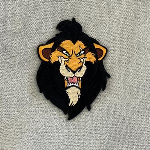 May include: Embroidered patch featuring a stylized lion's head with a black mane and a yellow face. The lion has a fierce expression with green eyes and an open mouth, revealing teeth. The patch is likely for clothing or accessories.