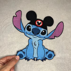 May include: A blue embroidered patch featuring the character Stitch from Lilo & Stitch. Stitch is wearing a black hat with Mickey Mouse ears and a red and white emblem. The patch has a light blue body, purple ears, and black eyes.