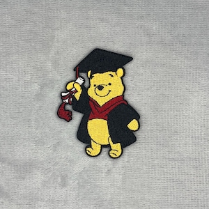 Puede incluir: Un parche bordado que muestra a un oso amarillo con un birrete negro y una toga negra con cuello rojo. El oso sostiene en su mano derecha un diploma blanco atado con una cinta roja, todo ello realizado con hilo de bordar detallado.