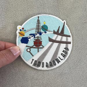 Puede incluir: Parche bordado con un diseño de Tomorrowland sobre un fondo de cielo azul claro. El parche incluye un cohete, planetas y la palabra "Tomorrowland" en blanco. El parche circular tiene un borde azul claro.
