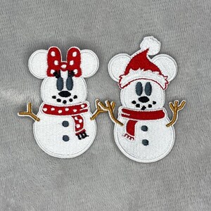 Op de afbeelding: Twee witte sneeuwmanpatches met muizenoren, zwarte ogen en een zwarte neus. Eén heeft een rode strik, de andere een rode kerstmuts. Beide hebben rode sjaals en takkenarmen. De patches zijn voor vakantieknutselwerk.