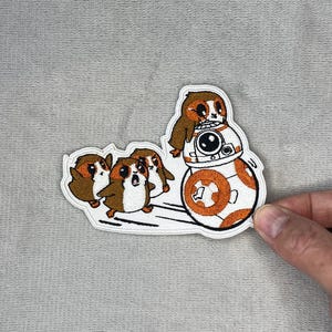 May include: Embroidered patch featuring Star Wars characters. The patch depicts three Porgs, a sloth riding on BB-8, and is outlined in white. The characters are in shades of brown, orange, and white.