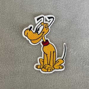 Op de afbeelding: Een geborduurde cartoon-hondpatch, geel met zwarte ogen, een rode halsband en een witte omtrek. De hond zit met zijn hoofd schuin.