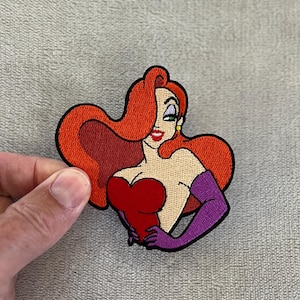 May include: Embroidered patch depicting a cartoon woman with vibrant red hair, a red dress, and purple gloves. The patch has a black border and is held by a hand against a neutral background. The woman has a seductive expression.